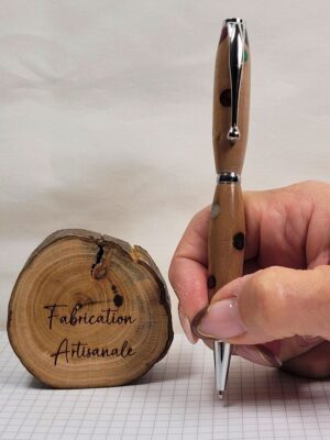 Stylo bille artisanal en bois d'alisier décoré de pois colorés incrustés dans la résine, tenu en main en position d'écriture. Les finitions chromées brillantes mettent en valeur le contraste entre le veinage naturel du bois et les motifs multicolores réalisés à la main.