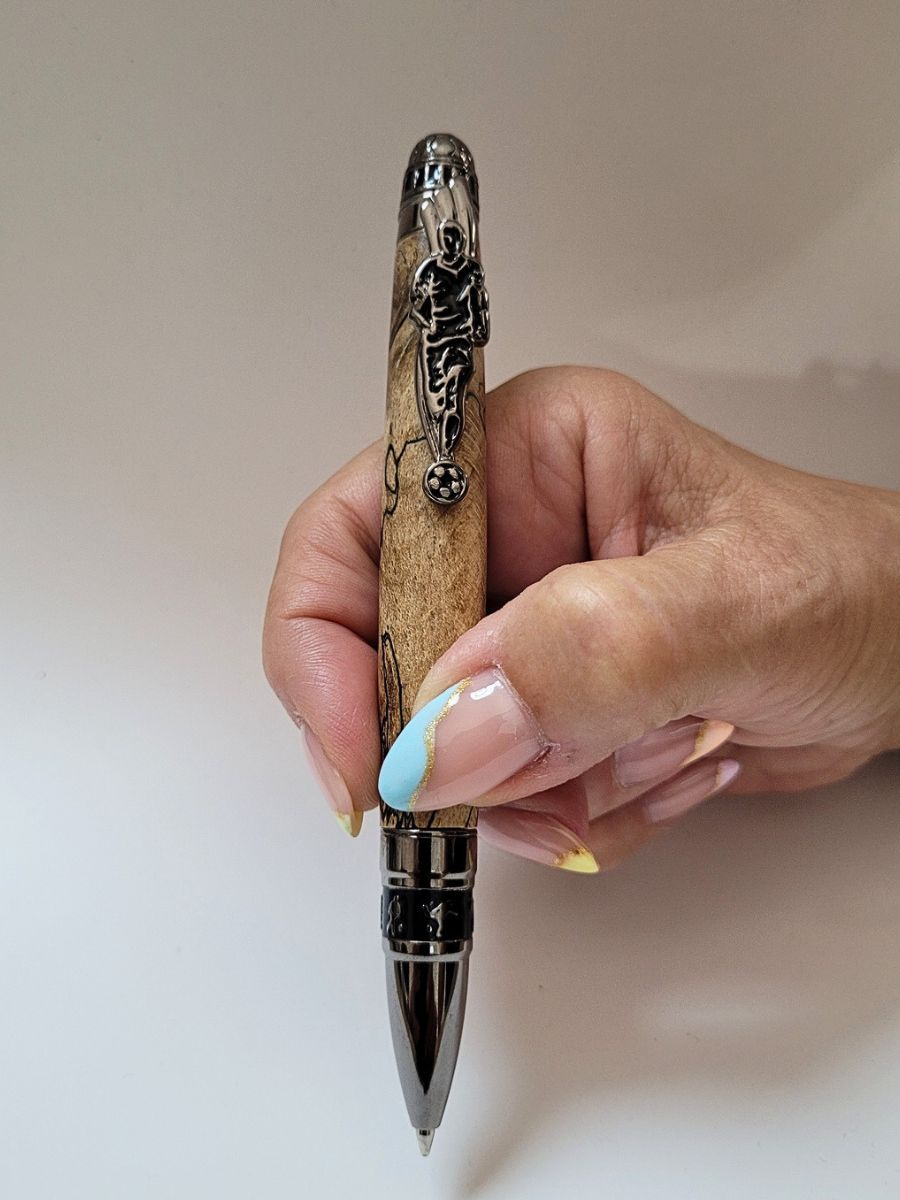 Stylo bille artisanal Football en hêtre échauffé tenu en main, mécanisme noir et argent avec joueur sculpté et ballon.
