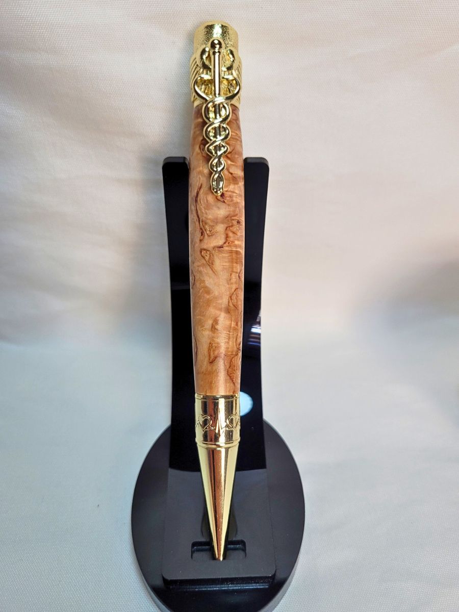 "Stylo médical en loupe de bouleau présenté verticalement sur support noir, clip caducée doré et pointe gravée de battements de cœur."