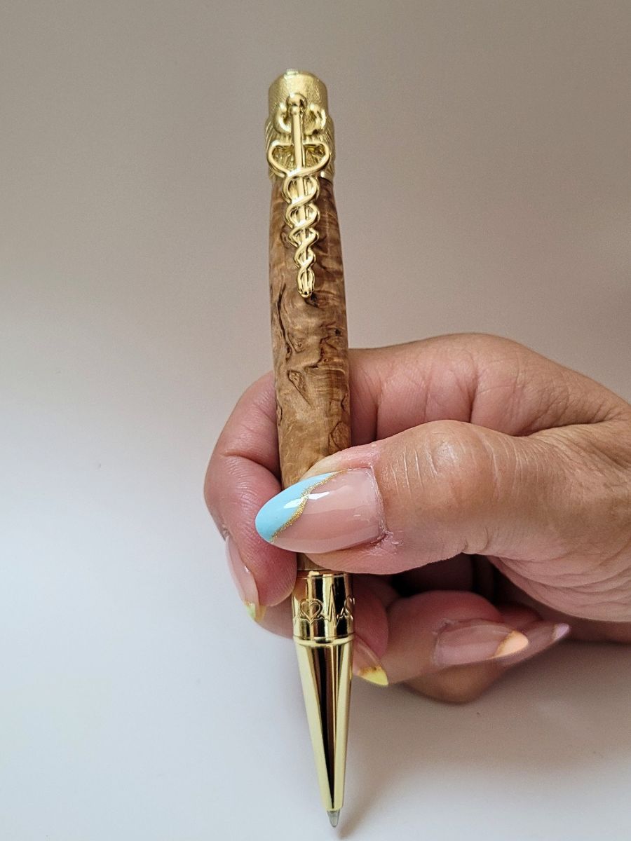 "Stylo médical en loupe de bouleau tenu en main, clip caducée doré et pointe gravée de battements de cœur, finition artisanale."