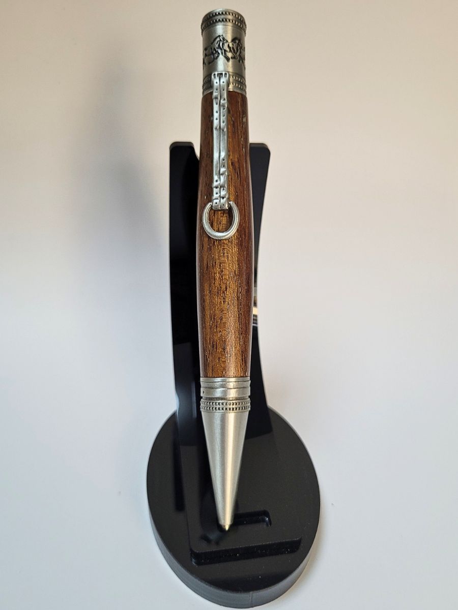 Stylo artisanal en chêne doré avec mécanisme en étain gravé de chevaux au galop, clip en forme d’étrier, présenté sur support noir.
