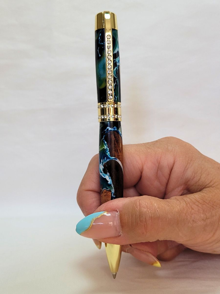 Stylo artisanal en résine époxy bleu et blanche avec bois de hêtre, finition dorée et strass, tenu en main.