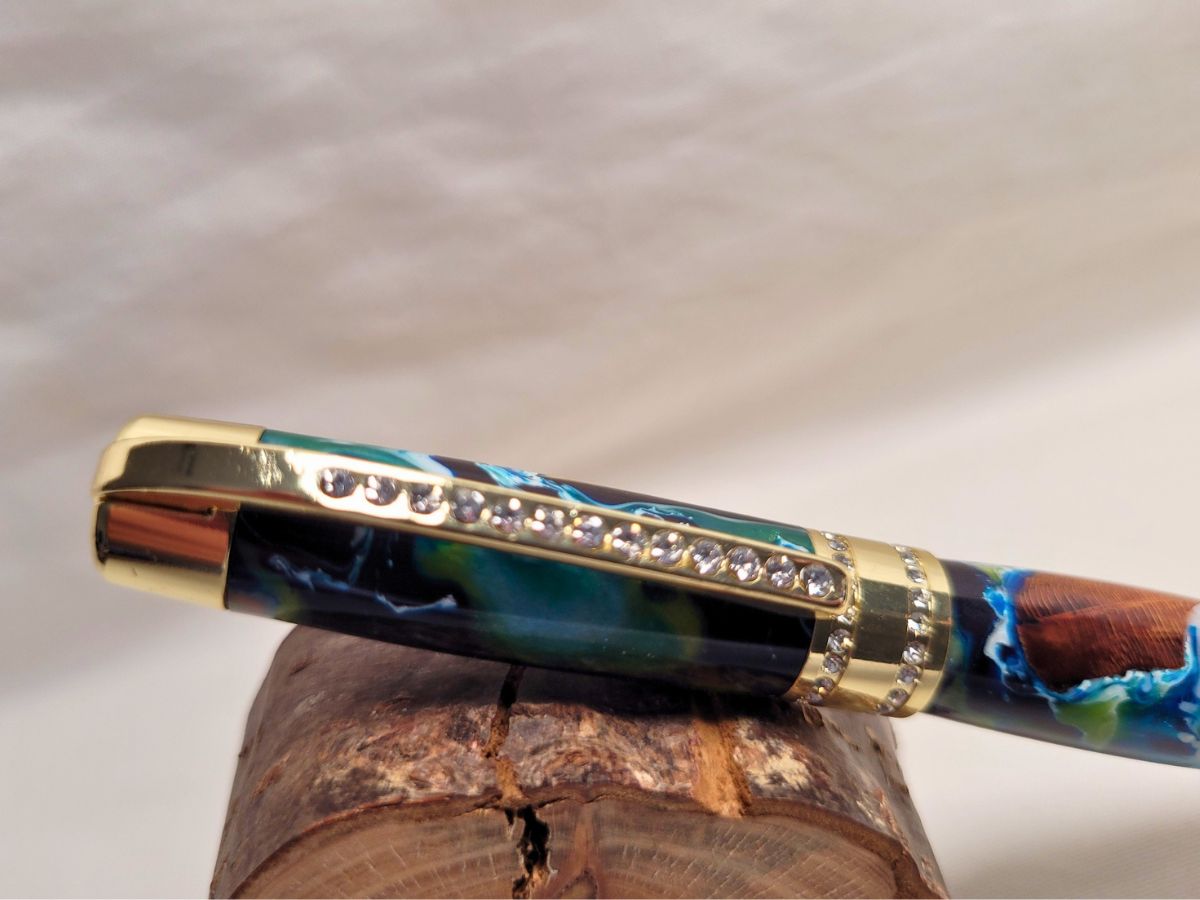 Stylo artisanal hybride en résine époxy bleu et blanche avec bois de hêtre incrusté, finition dorée et strass blancs, détail du capuchon.