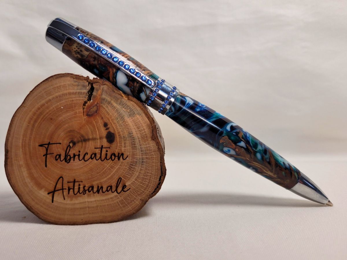 Stylo hybride artisanal en pomme de pin et résine époxy bleu-blanc-émeraude, finition chrome et strass bleus, posé sur rondin gravé Fabrication artisanale.