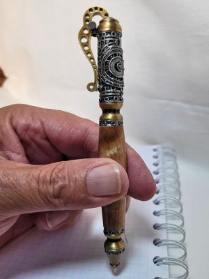 "Stylo steampunk artisanal en bois de bouleau, finitions étain et laiton, tenu en main au-dessus d’un carnet à spirale"