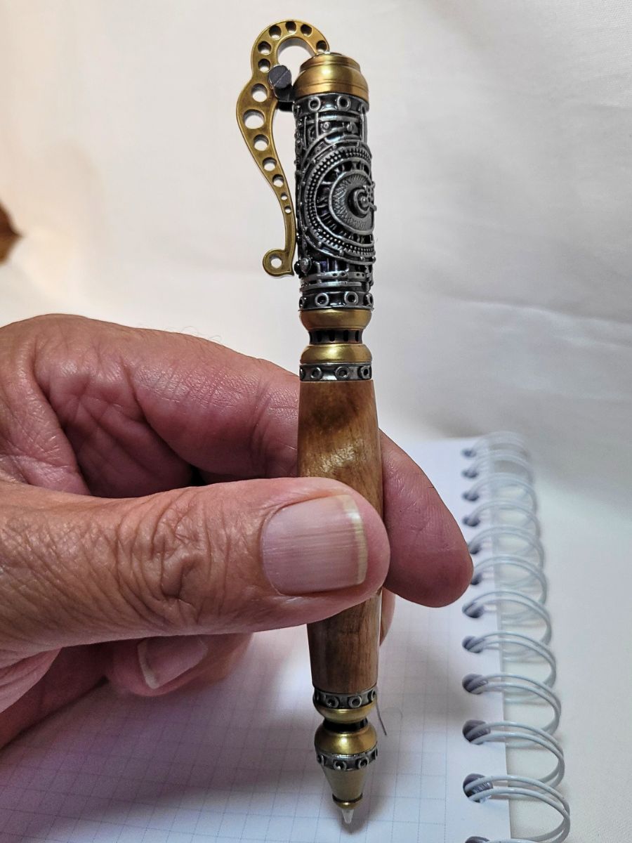"Stylo steampunk artisanal en bois de bouleau, finitions étain et laiton, tenu en main au-dessus d’un carnet à spirale"