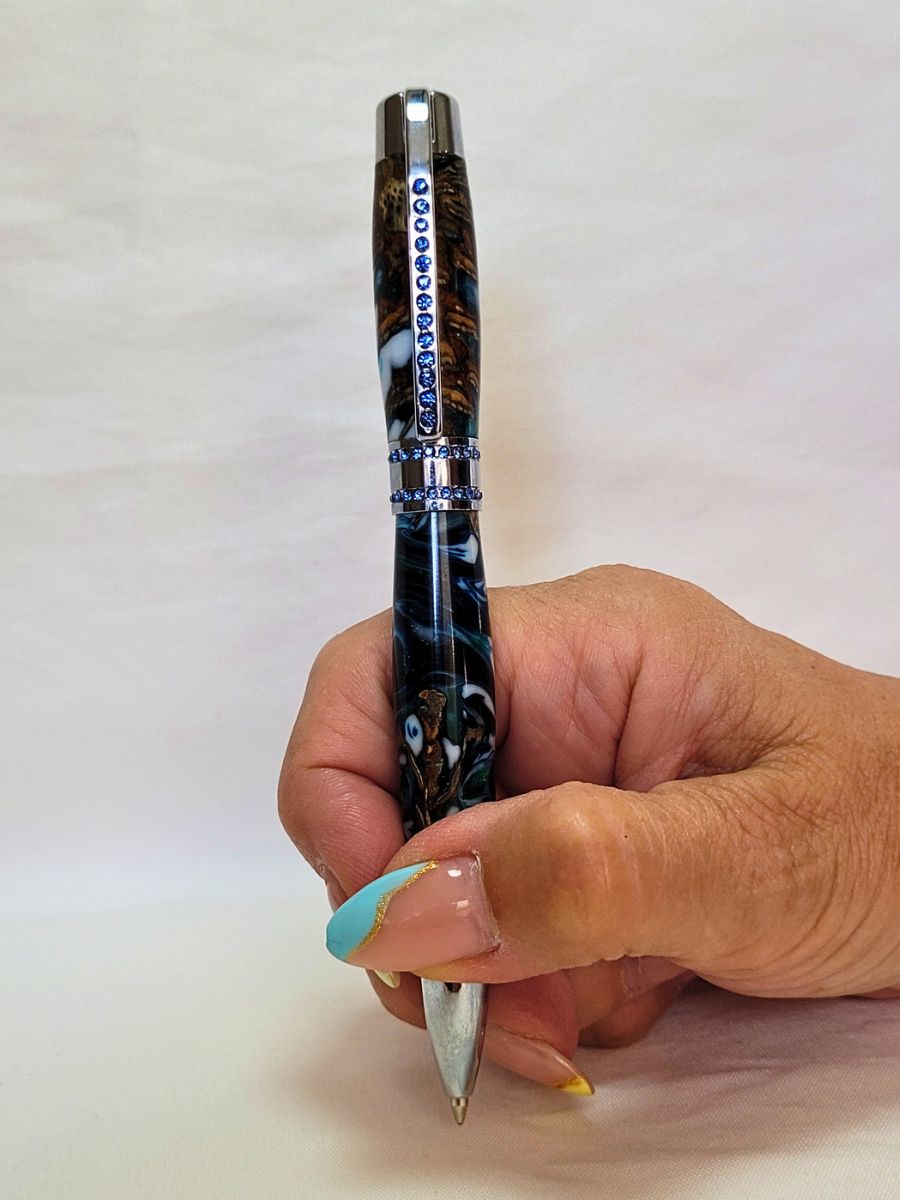 Stylo Princesse hybride en résine époxy et pomme de pin, teintes bleu profond et blanc nacré, finition chrome et strass bleus, tenu en main.