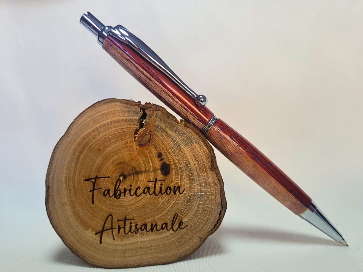 Porte‑mine artisanal en bois de padouk, frêne et bouleau, finition chromée, posé sur rondin gravé « Fabrication artisanale ».