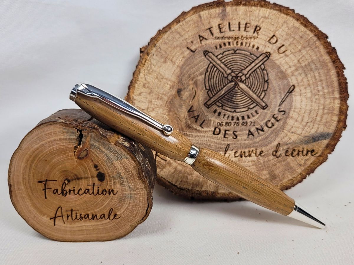 Stylo slim en bois de chêne massif, finition chrome brillant, posé sur rondin gravé Fabrication artisanale, logo L’Atelier du Val des Anges.