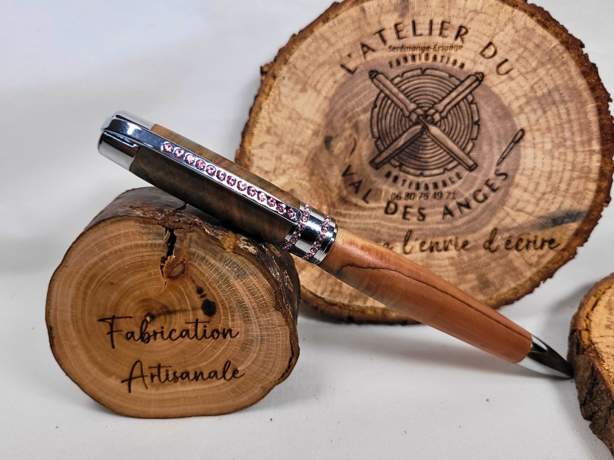 Stylo bille Princesse en bois de genévrier avec finition chromée et strass roses, présenté sur des rondins de bois dans un décor d'atelier artisanal français.