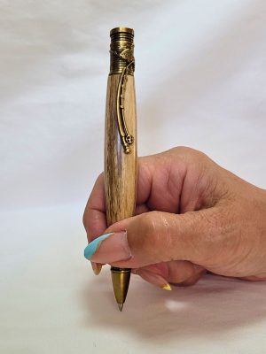 Stylo bille en bois de frêne avec placage bronze, tenu en main pour écrire. Clip canne à pêche et bague poisson ciselé visibles.