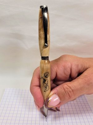 Stylo slim en bois de bouleau avec gravure moto, tenu en main lors de l’écriture sur papier quadrillé.