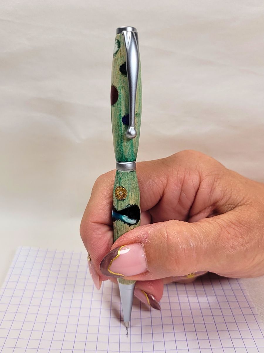 Photo montrant le stylo artisanal en bois teinté vert à pois multicolores pris en main pour démontrer son confort d'écriture sur un papier à carreaux.