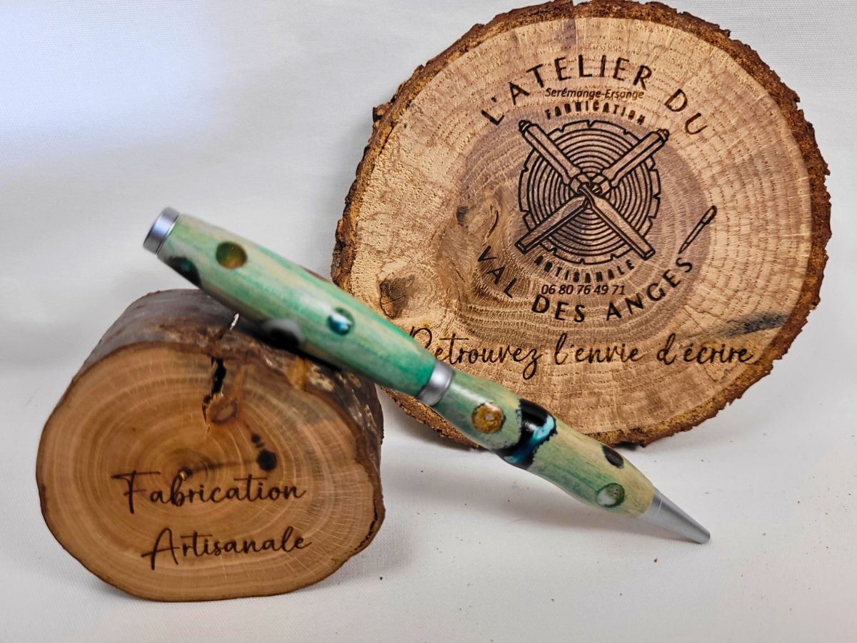Photo montrant un stylo artisanal en bois teinté vert à pois multicolores, présenté sur un rondin de bois gravé devant le logo de L'Atelier du Val des Anges.