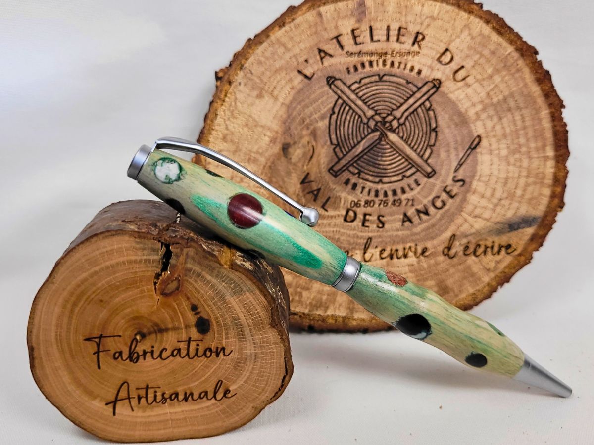 Photo montrant le stylo artisanal en bois teinté vert à pois multicolores présenté en équilibre sur un rondin de bois devant le logo de L'Atelier du Val des Anges.