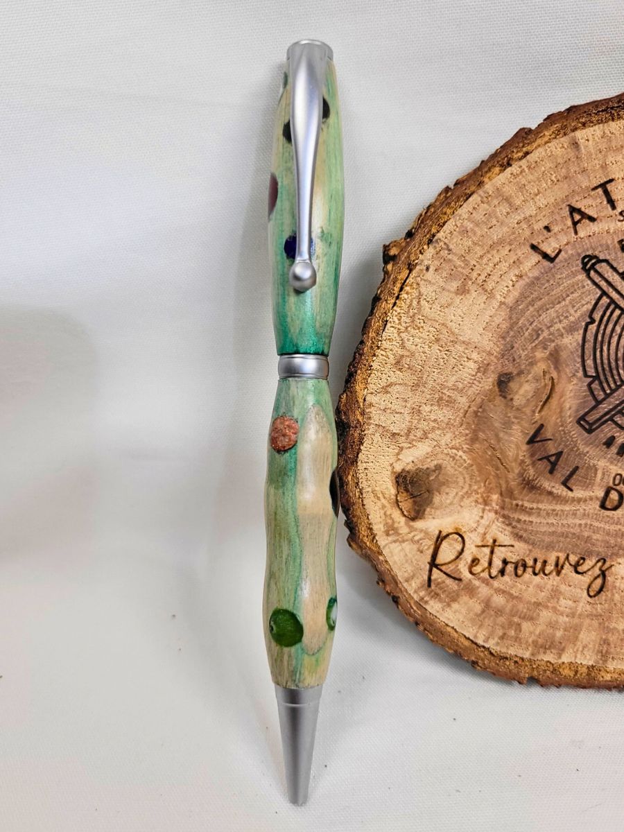 Photo montrant un stylo artisanal en bois teinté vert avec inclusions de couleurs multicolores, présenté verticalement devant un rondin gravé de L'Atelier du Val des Anges.