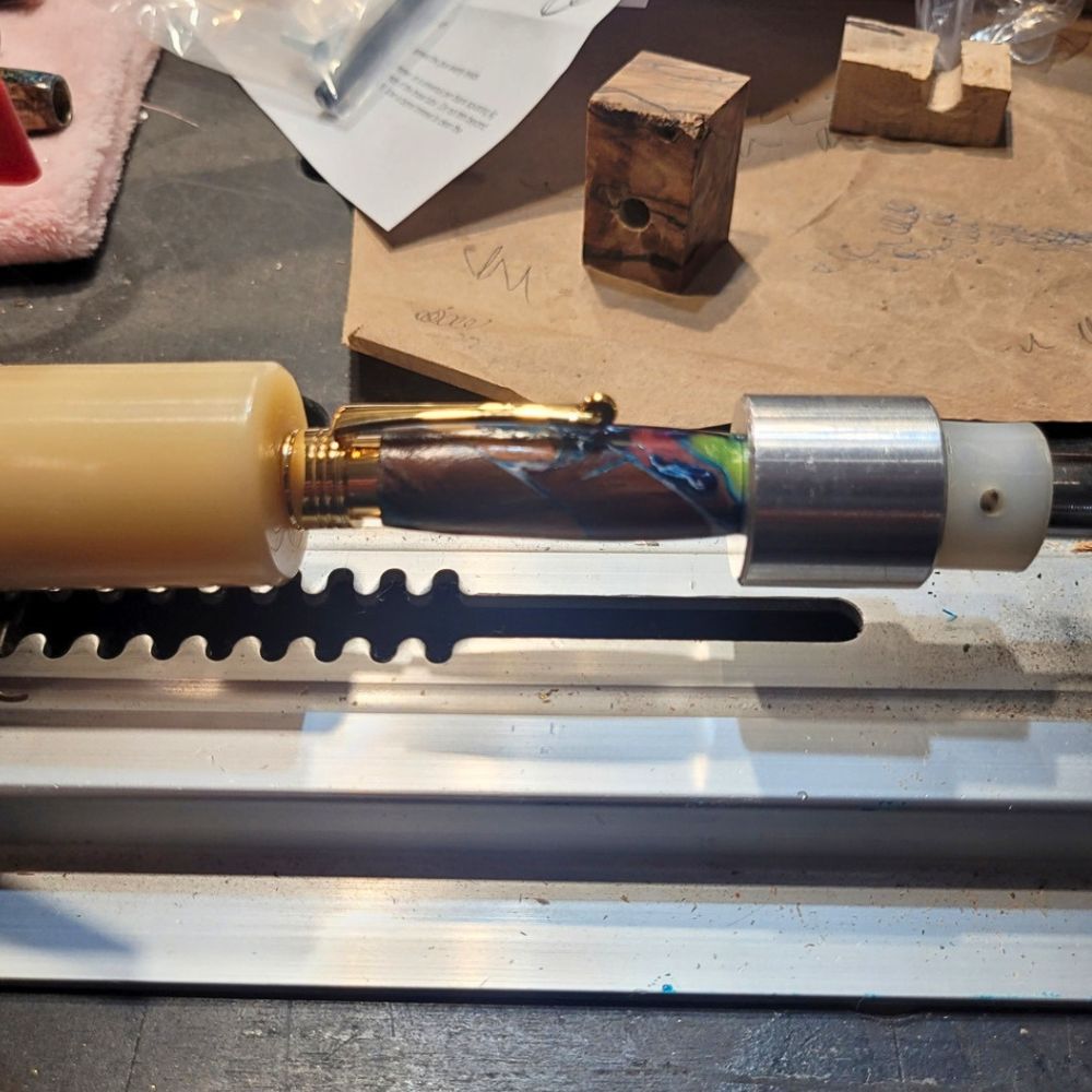 Assemblage d'un stylo artisanal sur tour à bois dans l'atelier du Val des Anges, corps en résine aux nuances bleues, vertes et cuivrées monté sur un mandrin de précision, avec mécanisme métallique doré en cours d'ajustement avant le tournage et la finition manuelle