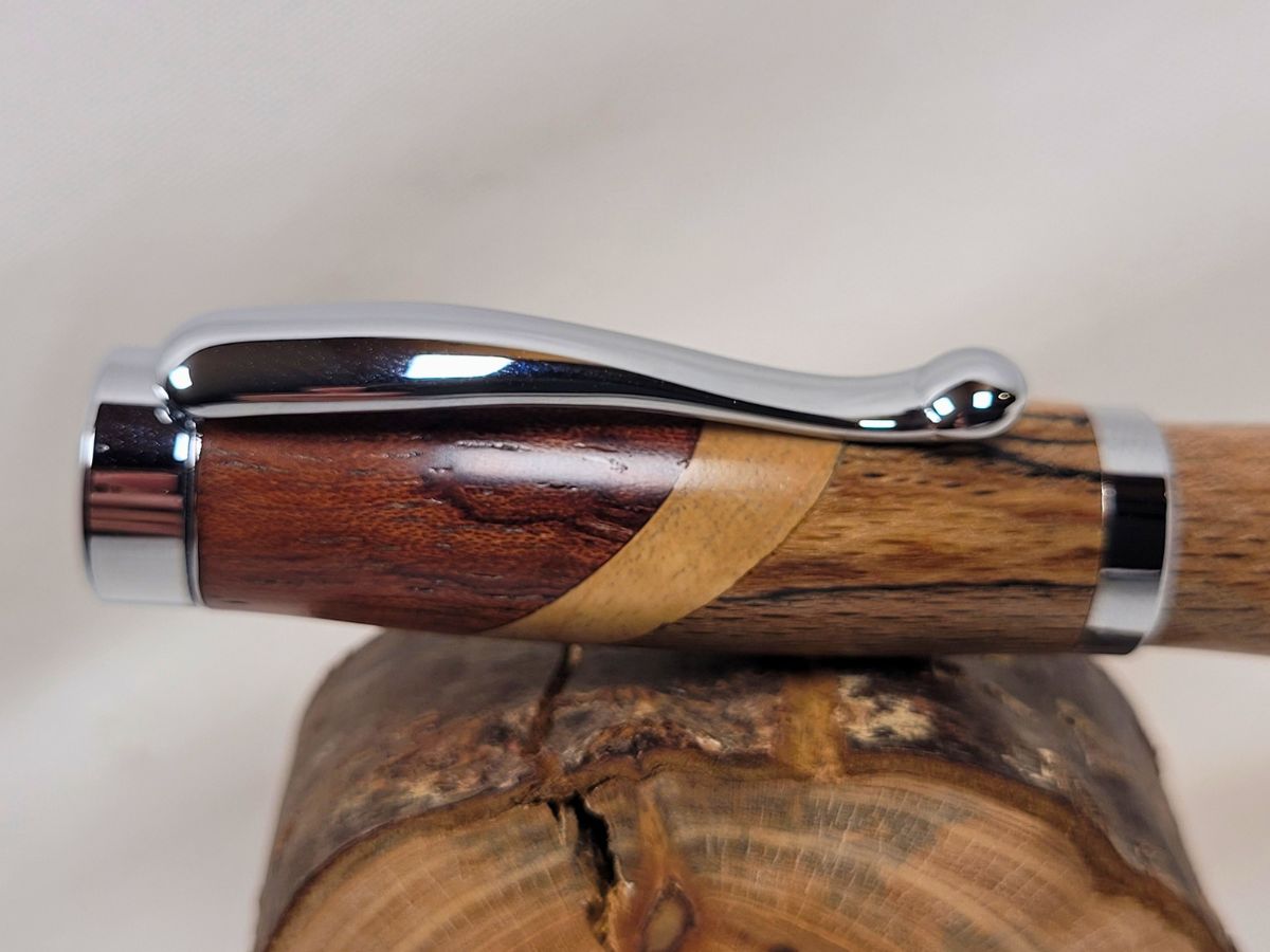 Gros plan sur le capuchon chromé brillant d'un roller artisanal en assemblage de hêtre échauffé, bouleau et padouk, mettant en valeur les contrastes naturels entre les différentes essences de bois.