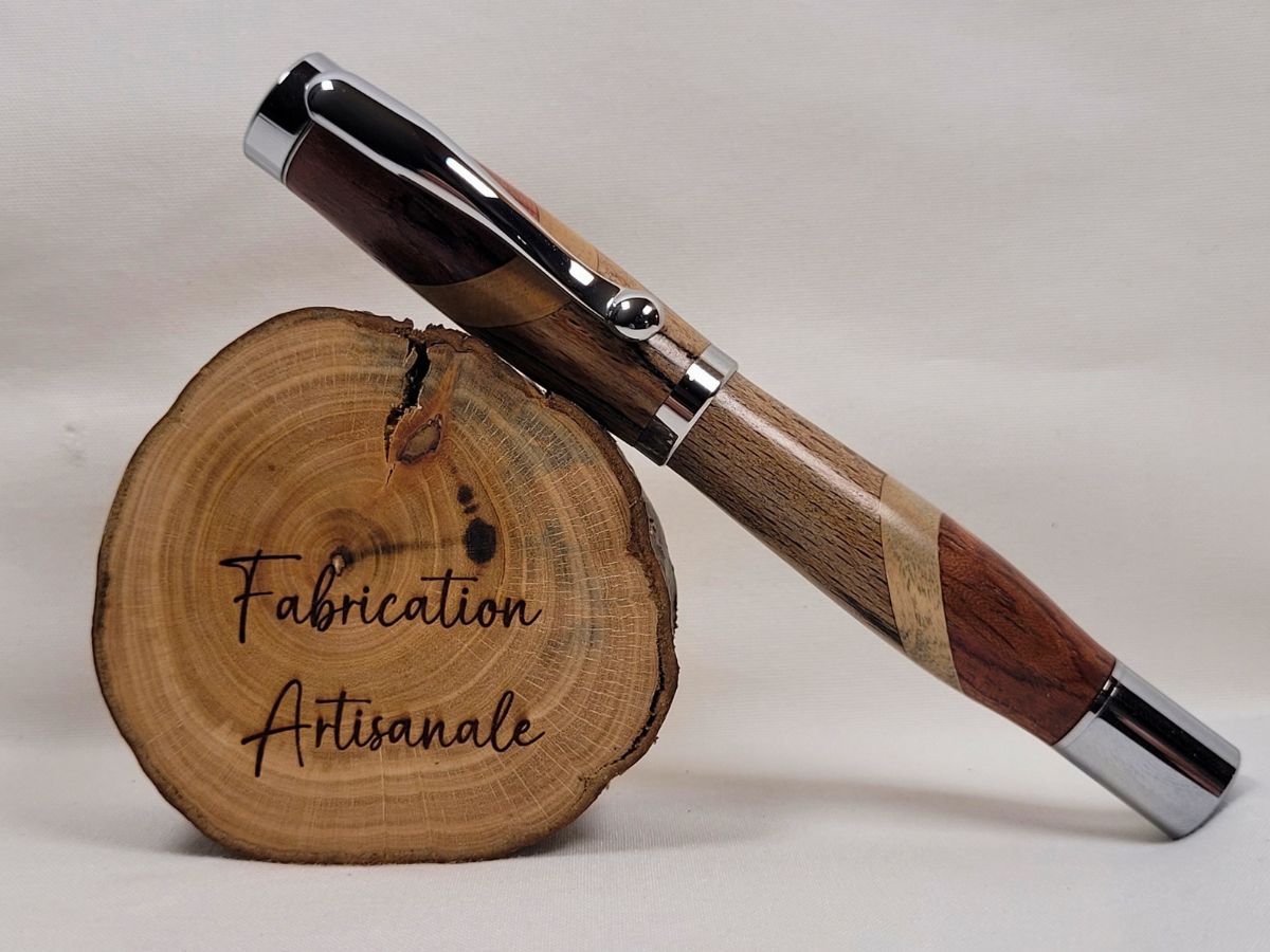 Stylo roller artisanal fermé en assemblage de hêtre échauffé, bouleau et padouk avec placage chromé brillant, posé sur une rondelle de bois, mettant en valeur les contrastes naturels entre les différentes essences de bois.