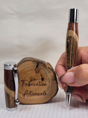 Roller artisanal en assemblage de hêtre échauffé, bouleau et padouk tenu en main, finition chromée brillante, présenté avec son capuchon devant une rondelle de bois gravée « Fabrication Artisanale », création unique réalisée à la main en Moselle.
