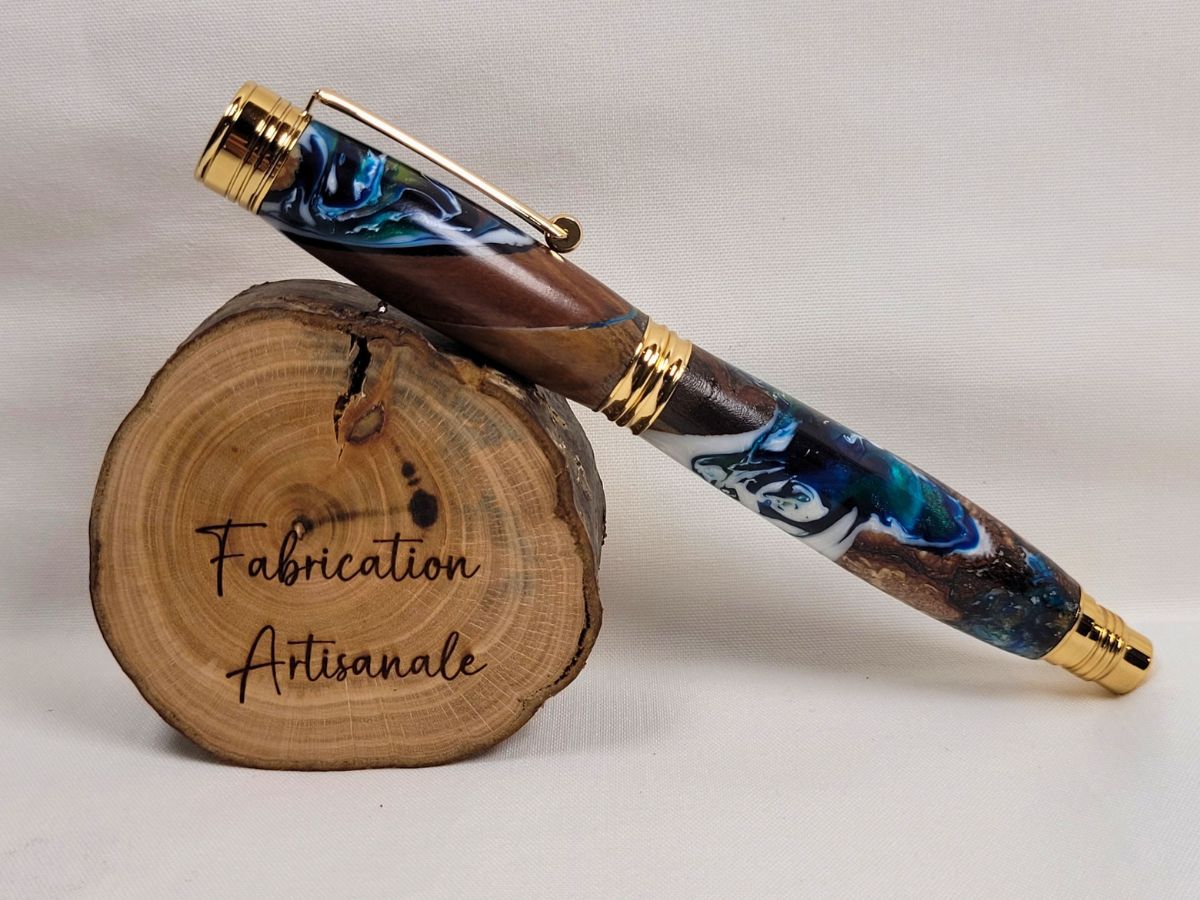 Stylo roller artisanal en bois et résine époxy bleu blanc vert avec placage doré, présenté sur un rondin de bois, pièce unique fabriquée à la main dans mon atelier en Moselle