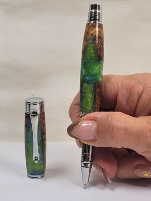 Stylo roller artisanal en racine de hêtre et résine époxy aux effets marbrés verts et bleus, tenu en main pour illustrer ses dimensions, équipé d'une cartouche roller à bille céramique pour une écriture fluide