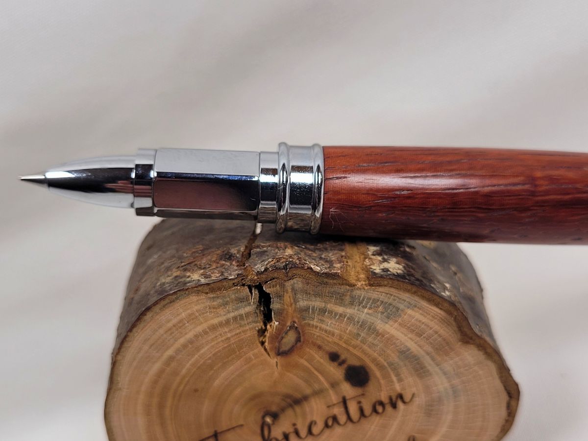 Gros plan sur la pointe d'écriture d'un stylo roller artisanal en bois de padouk avec finition chromée. Les veinures rouge brun du padouk sont visibles sur le corps du stylo poli à la main. Fabrication artisanale française réalisée dans l'atelier du Val des Anges.