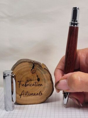Stylo roller artisanal en bois de padouk tenu en main, avec capuchon hexagonal magnétique chromé posé à proximité, démonstration de la prise en main et de l'équilibre du stylo.
