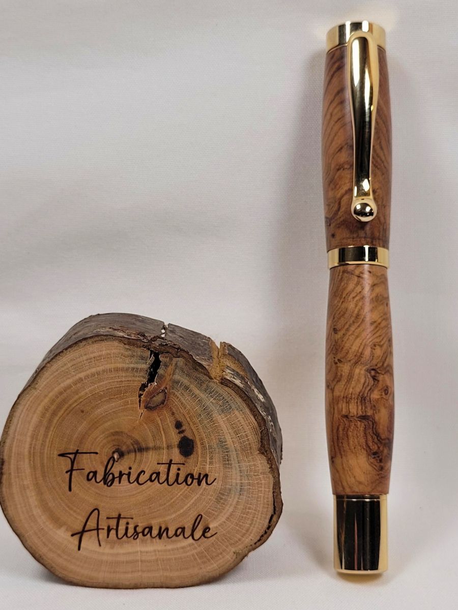 Roller artisanal en bois d'olivier fermé avec finition plaquée or brillante, présenté debout devant une rondelle de bois gravée « Fabrication Artisanale ». Les veinages naturels et les contrastes du bois soulignent le caractère unique de cette création artisanale française.Stylo roller artisanal en bois d'olivier avec placage or brillant, création française haut de gamme présentée fermée, mettant en valeur les nuances naturelles du bois et un savoir-faire artisanal traditionnel.