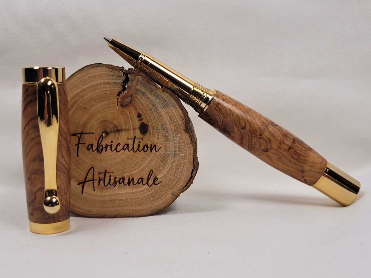 Roller artisanal en bois d'olivier ouvert avec placage or brillant, présenté devant une rondelle de bois gravée « Fabrication Artisanale ». Les veinages tourmentés et les nuances chaleureuses du bois d'olivier mettent en valeur cette pièce unique réalisée à la main en France. Recharge roller à pointe céramique pour une écriture fluide et précise.