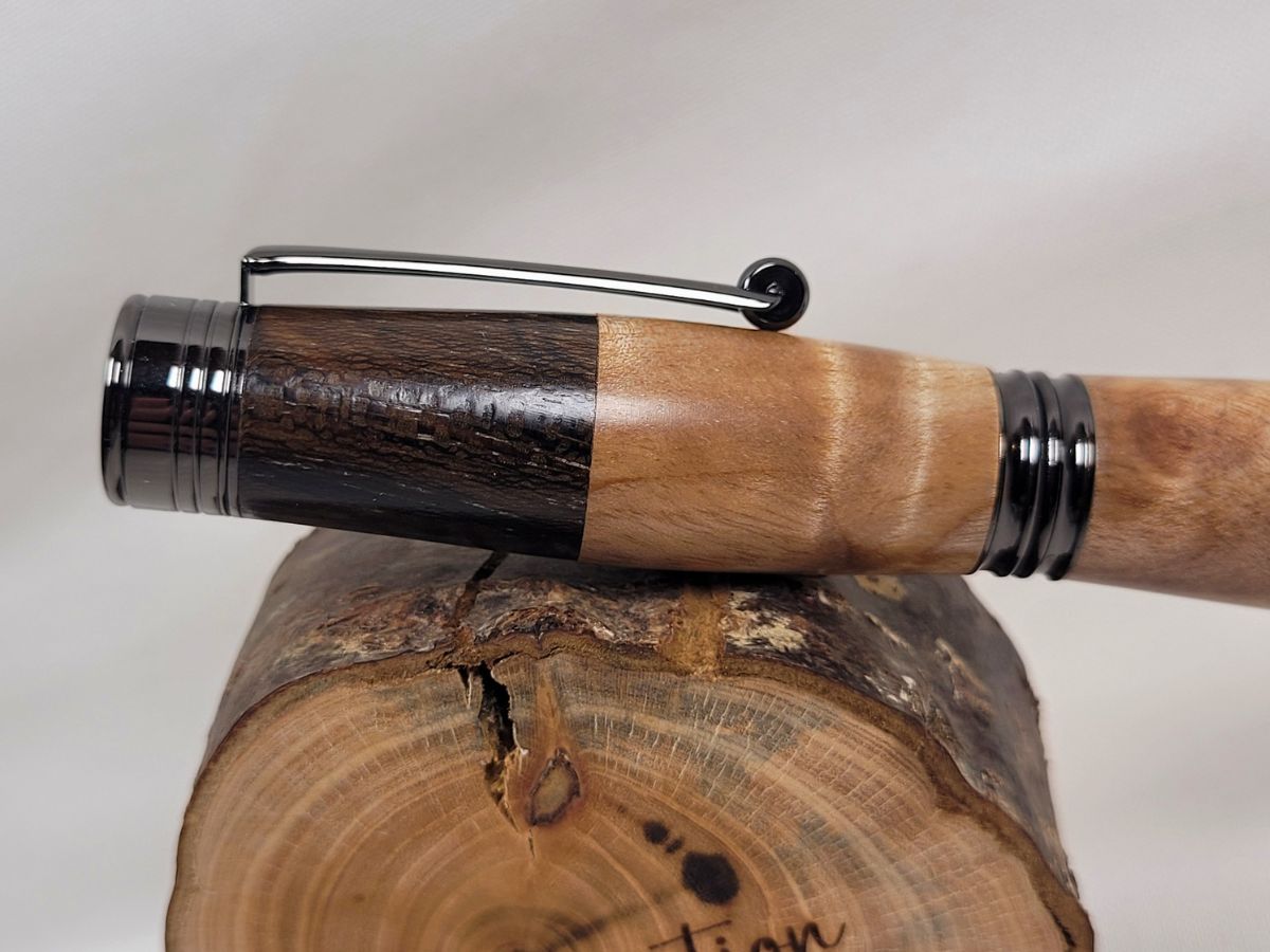 Gros plan sur le capuchon d'un stylo roller artisanal en bouleau ondé et palissandre. La jonction entre les deux essences met en évidence le contraste entre le bois clair aux reflets satinés du bouleau et les teintes plus sombres du palissandre. L'épingle et les finitions en placage noir brillant renforcent l'élégance de cette pièce unique façonnée à la main.
