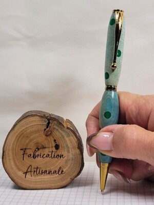 Stylo slim artisanal en charme teinté bleu avec incrustations vertes en résine époxy tenu en main pour illustrer ses dimensions, placage doré, clip à épingle boule et fabrication artisanale française