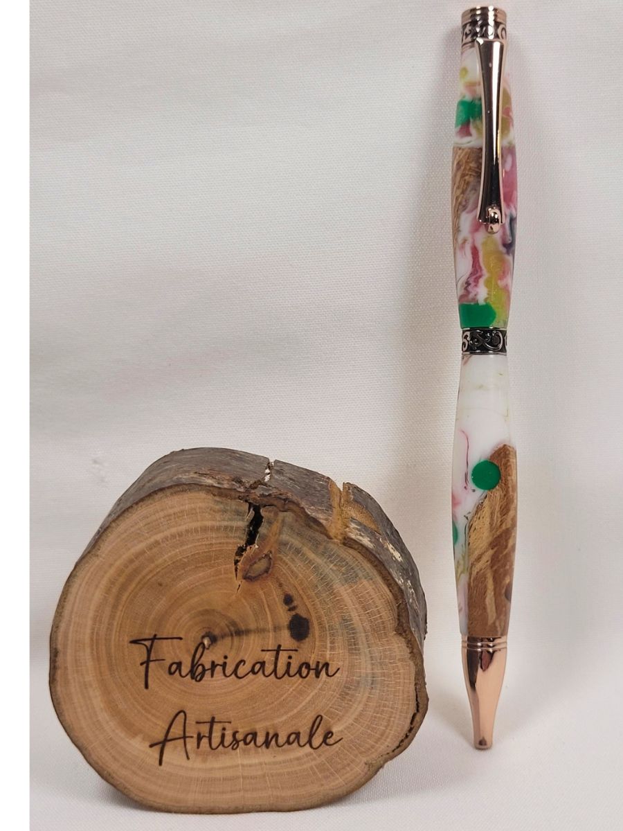 Stylo slim premium artisanal en bois de hêtre et résine époxy multicolore, finition or rose, bagues décoratives finement ciselées, motifs marbrés rose blanc vert avec pois colorés, présenté à côté d'un rondin marqué "Fabrication Artisanale"