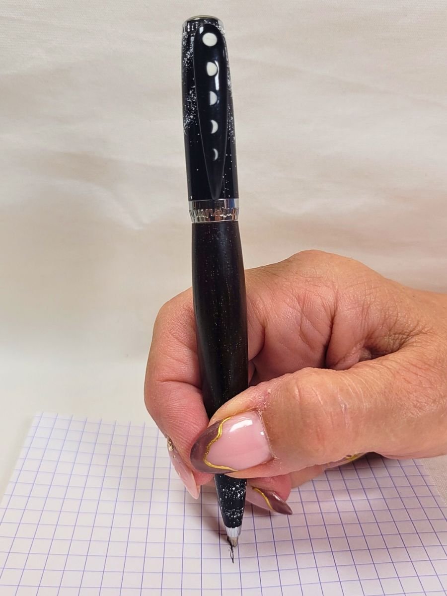 Détail d'utilisation d'un stylo bille artisanal en ébène décoration espace et galaxie, tenue en main lors de l'écriture, finitions chromées et motif phases lunaires