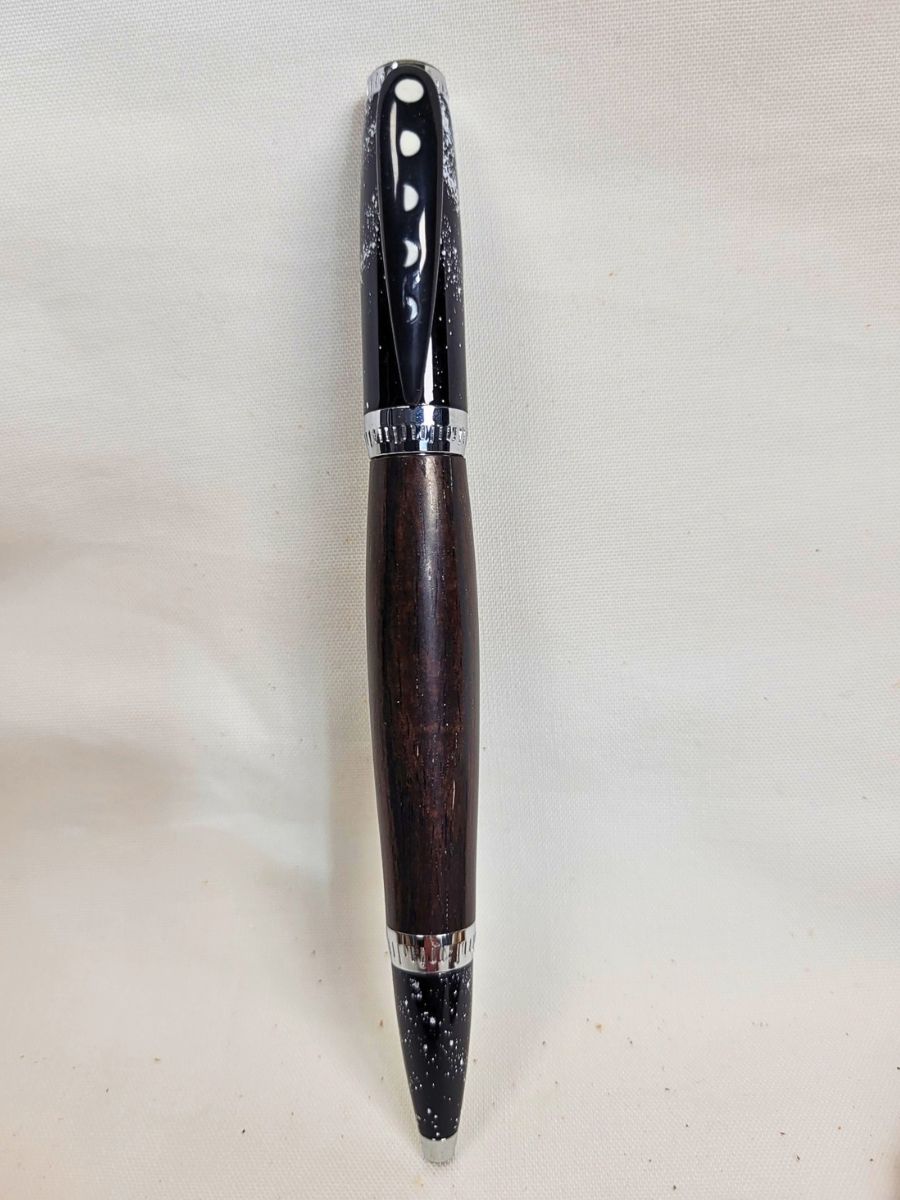 Stylo bille galaxie en bois d'ébène présenté debout sur fond clair, finitions chromées, pointe étoilée et épingle représentant les différentes phases de la lune