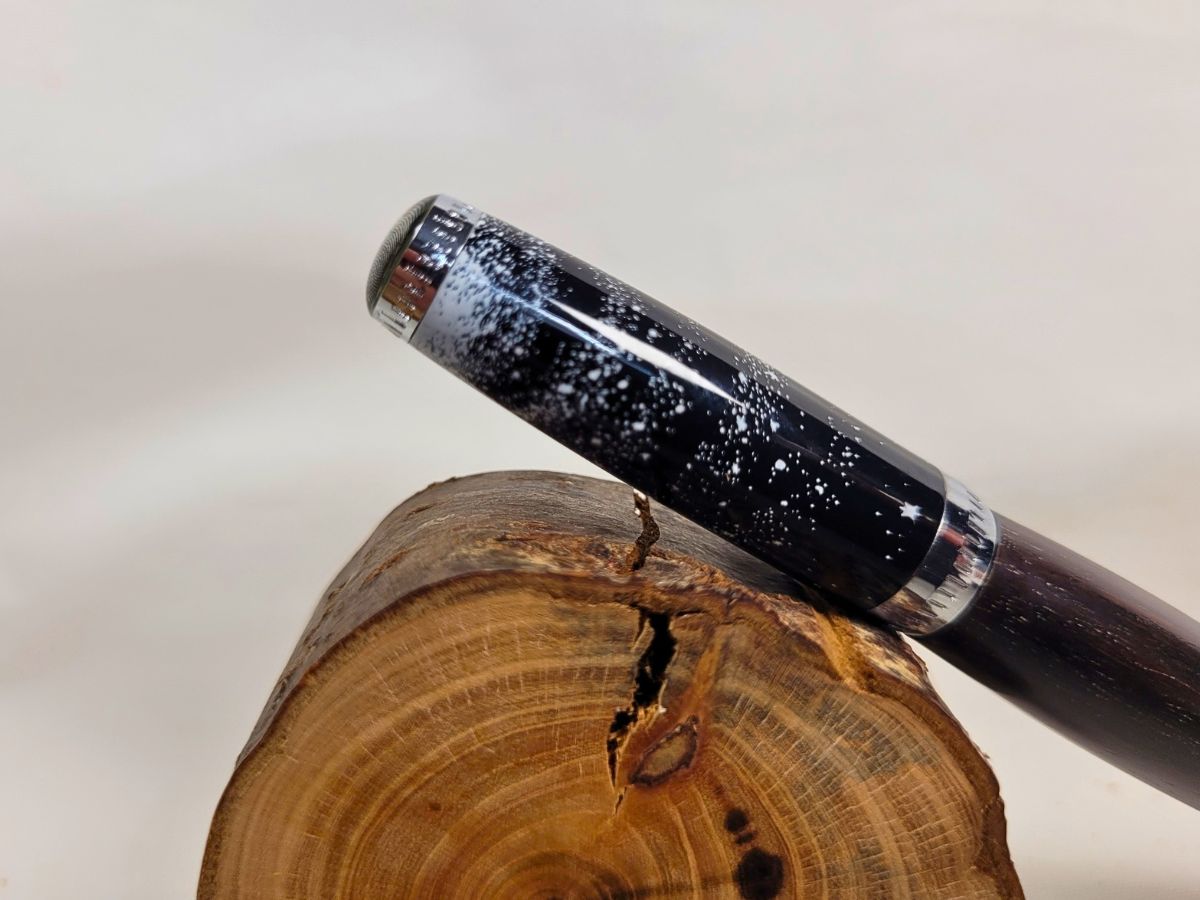 Détail du capuchon d'un stylo bille en ébène tourné à la main, finition chromée et motif galaxie noir parsemé d'étoiles blanches évoquant l'espace et l'univers, création artisanale française unique.