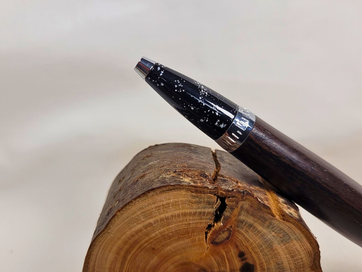 Gros plan sur la pointe d'un stylo bille artisanal en ébène avec finition chrome, décor galaxie noir constellé d'étoiles blanches rappelant la Voie lactée, fabrication française réalisée à la main.