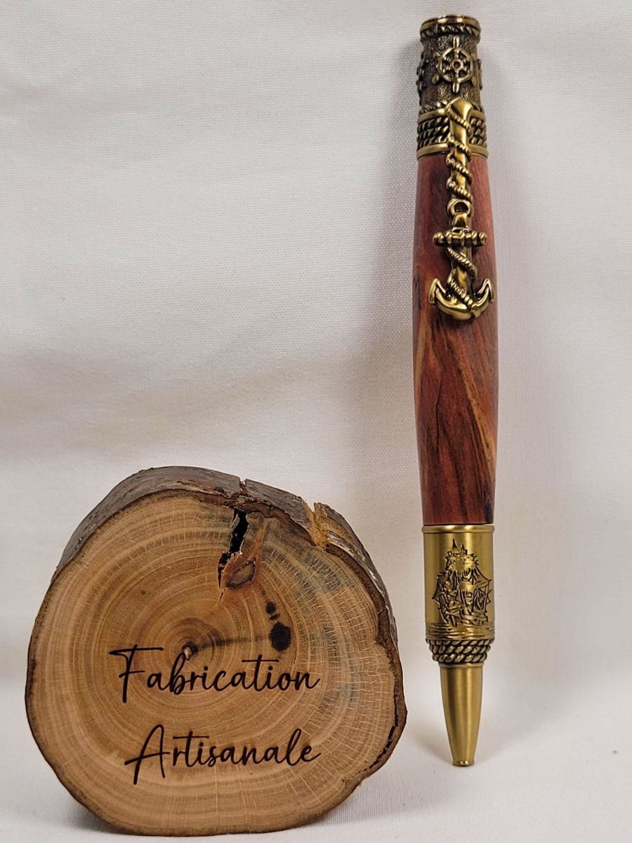 Stylo en bois de cerisier artisanal avec placage en laiton ciselé, ancre de bateau formant le clip, boussole décorative au sommet, voilier gravé sur la partie inférieure et support en rondin de bois marqué Fabrication Artisanale.