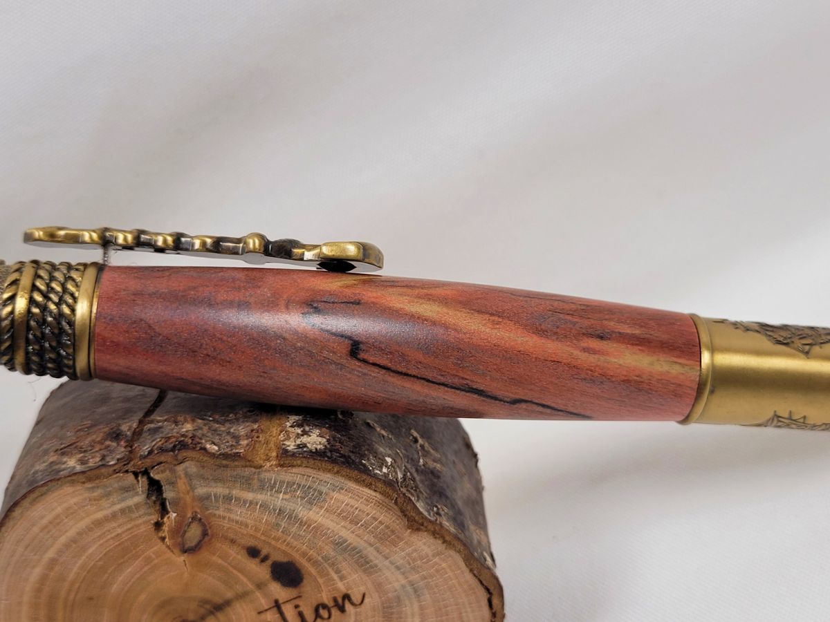 Gros plan sur le corps d'un stylo artisanal en bois de cerisier présentant un superbe veinage rouge et brun aux nuances naturelles, associé à des finitions en laiton ciselé sur le thème de la marine et du voyage maritime.