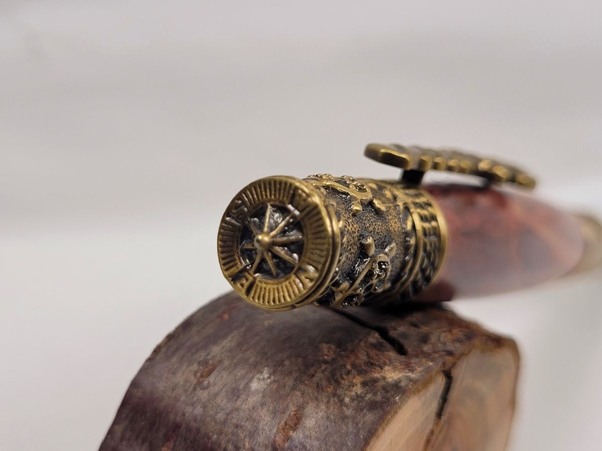Stylo artisanal en bois de cerisier tourné à la main avec gros plan sur la boussole en laiton ciselé située au sommet du capuchon, finitions marines haut de gamme, veinage rouge naturel du bois et décoration nautique inspirée de la navigation.