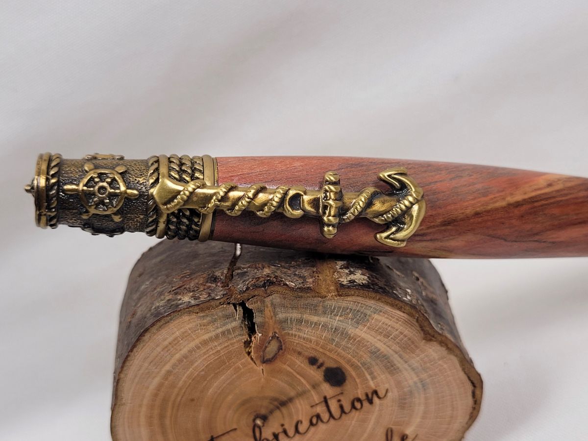 Détail du clip en forme d'ancre de bateau sur un stylo artisanal en bois de cerisier tourné à la main, avec finitions en laiton ciselé. Le veinage rouge et brun du cerisier contraste élégamment avec les ornements marins comprenant une ancre, un gouvernail et des cordages nautiques. Création artisanale française inspirée de l'univers de la navigation.