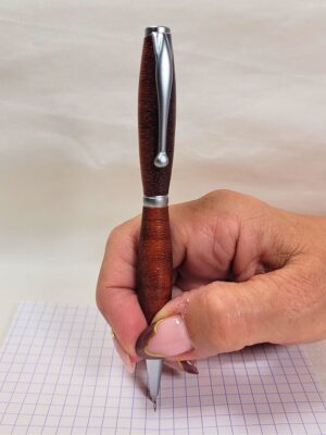 Stylo slim en padouk tenu en main en position d'écriture afin d'illustrer son ergonomie et son format fin. Les fibres traversantes créent un motif naturel unique mis en valeur par les finitions chrome satiné.