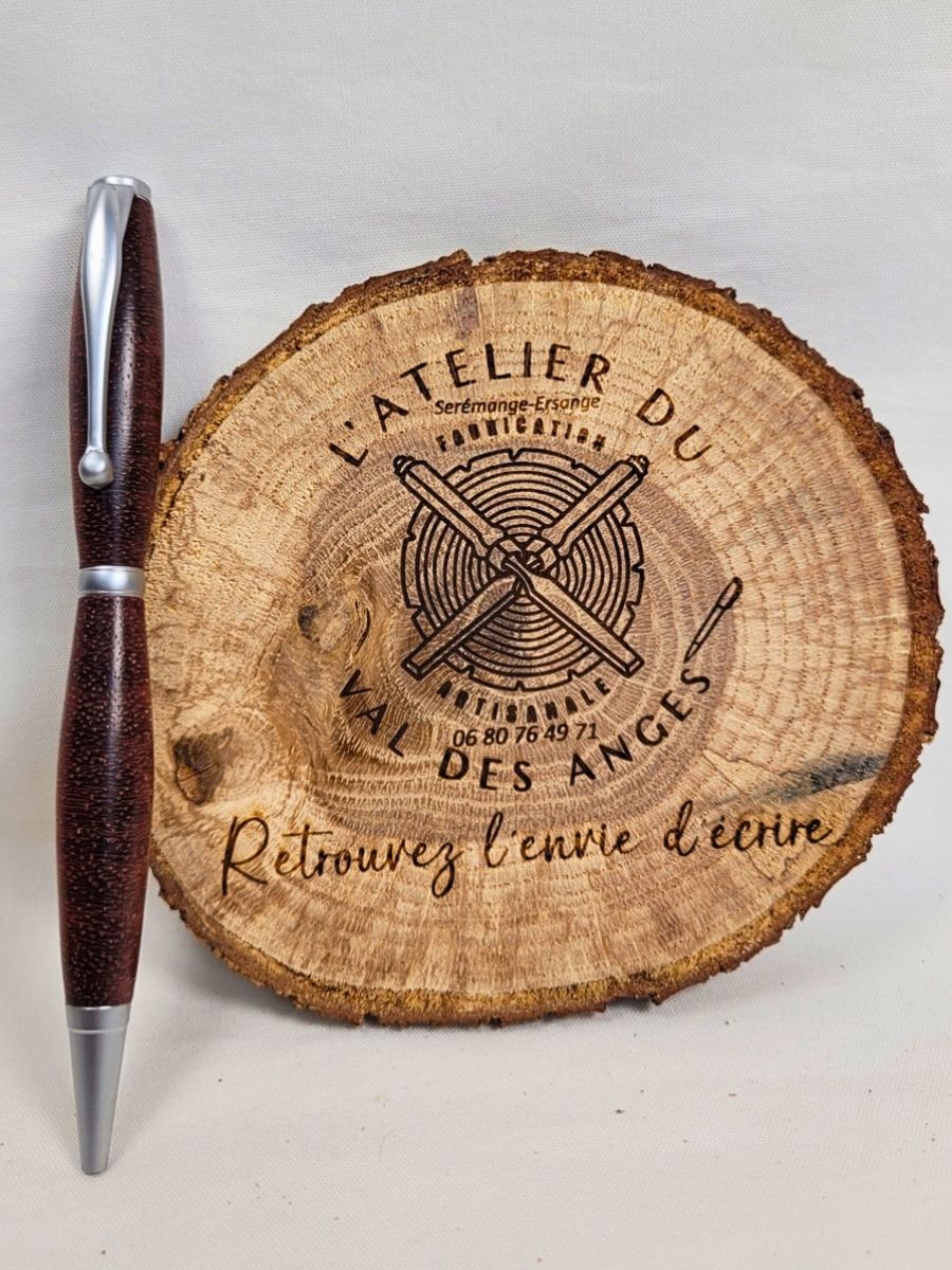 Vue complète d'un stylo artisanal en bois de padouk tourné dans les fibres de travers, placé devant le logo de l'Atelier du Val des Anges. Le contraste entre les teintes rouge acajou du bois et le chrome satiné souligne l'élégance de cette création unique.