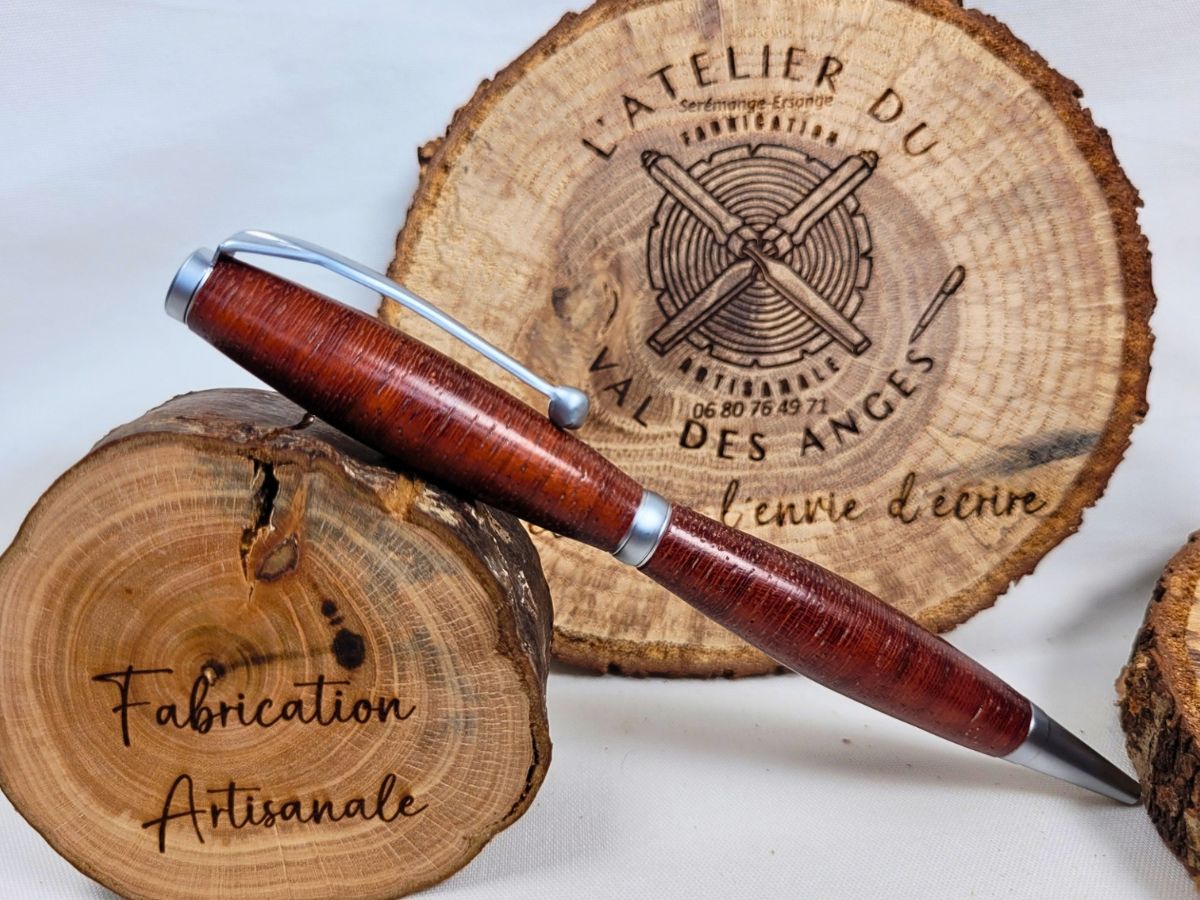 Stylo slim artisanal en bois de padouk présenté sur un support en bois gravé Fabrication Artisanale. Le veinage rouge brun du padouk tourné dans les fibres de travers contraste élégamment avec les attributs en chrome satiné et l'épingle à boule.