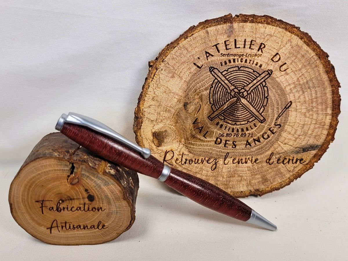 Stylo slim artisanal en bois de padouk tourné dans les fibres de travers, présenté sur un support en bois gravé « Fabrication Artisanale ». Les nuances rouge brun profond du padouk s'accordent harmonieusement avec les finitions chrome satiné et l'épingle à boule, mettant en valeur le caractère unique de cette création réalisée à la main.