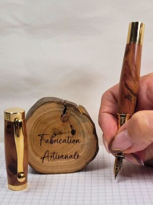 Stylo plume artisanal en genévrier avec plume dorée et pointe iridium tenu en position d'écriture, capuchon vissé, bois aux motifs tourmentés et nuances naturelles brun rougeâtre