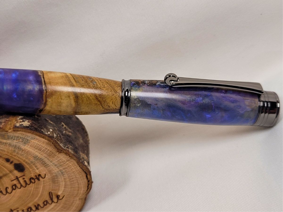 Gros plan sur le capuchon d'un stylo roller artisanal en résine époxy mauve et bois de hêtre. La résine présente des nuances violettes profondes avec des effets de lumière évoquant une nébuleuse. Le clip et les finitions noir brillant apportent une touche contemporaine à cette pièce unique fabriquée à la main.