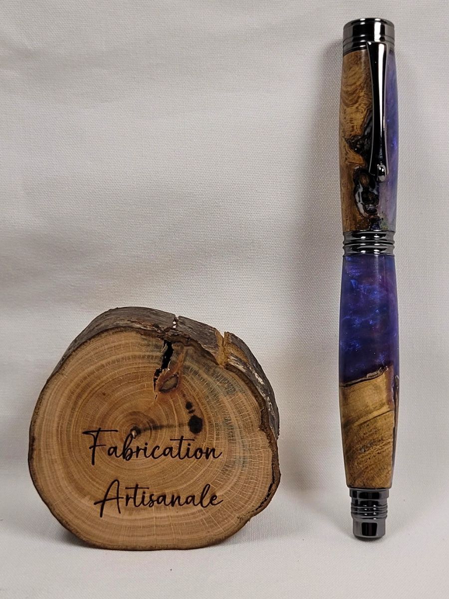 Stylo roller artisanal hybride en hêtre et résine époxy mauve aux reflets violets et bleutés, présenté fermé à côté d'un rondin de bois gravé « Fabrication Artisanale ». Les attributs noir brillant mettent en valeur le contraste entre le veinage naturel du hêtre et la profondeur translucide de la résine. Création unique réalisée à la main dans notre atelier.