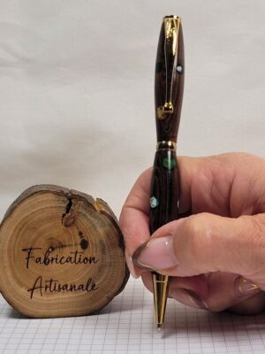 Stylo slim artisanal en palissandre avec incrustations de pois colorés en résine, finition dorée brillante, tenu en main pour illustrer la prise en main et les proportions du stylo, présenté à côté d'un rondin de bois gravé "Fabrication Artisanale", création unique réalisée à la main dans l'atelier du Val des Anges.